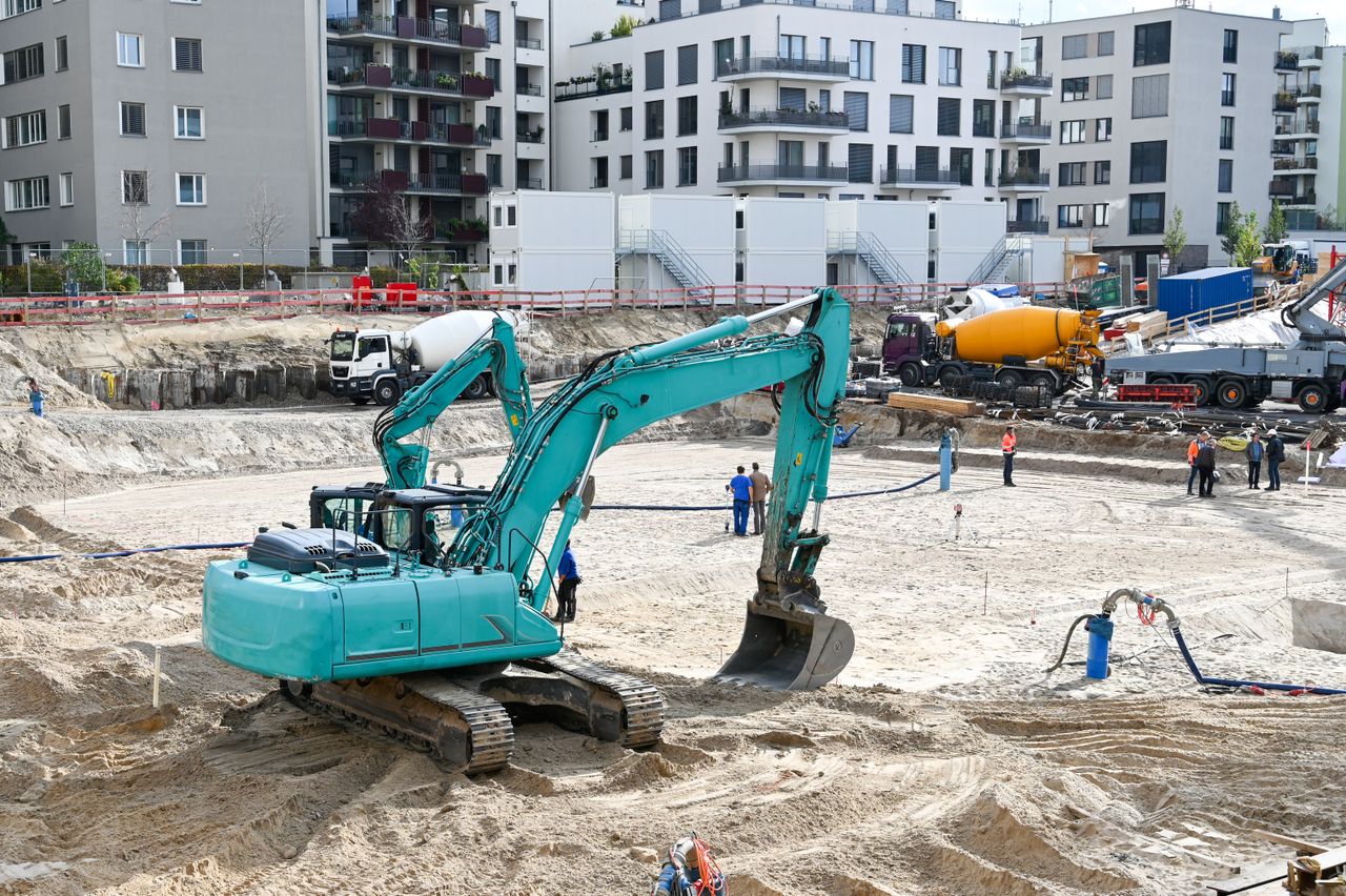 A busy construction site with machinery, workers, and residential buildings in the background.